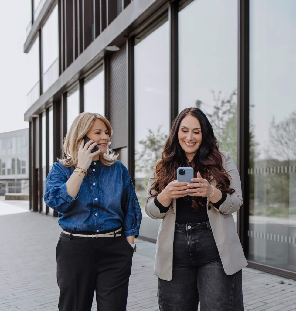 Zwei Frauen laufen neben einem Glasgebäude während die eine telefoniert und die anderen auf ihr Handy schaut