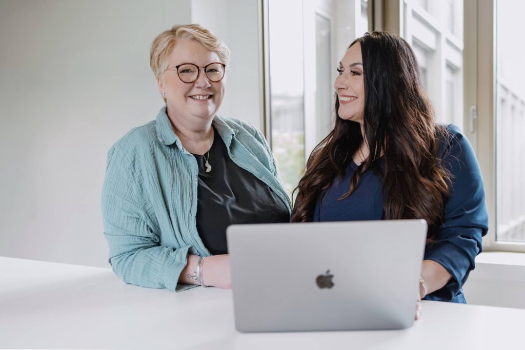 Zwei Mitarbeiterinnen von GIB21 stehen vor einem Macbook und führen ein Gespräch