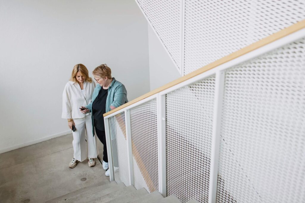 Zwei BEM Beraterinnen von GIB21 unterhalten sich auf einer Treppe während sie auf ein Smartphone schauen