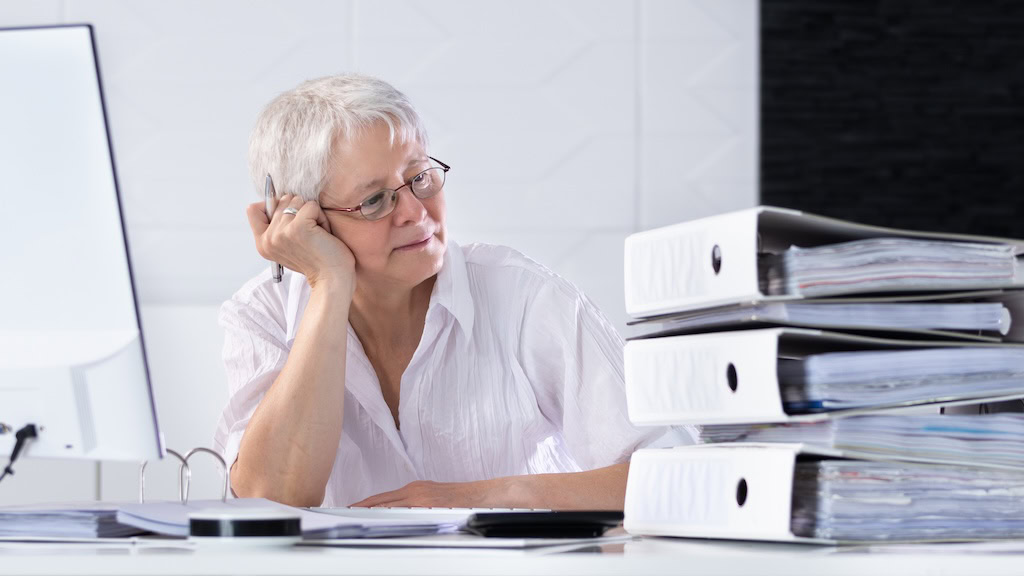 Ältere Frau arbeitet am Computer neben einem Stapel Aktenordner